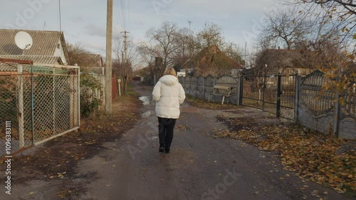 Young woman walking along a muddy dirt road past old and poor houses in a run-down area, wearing a warm jacket.