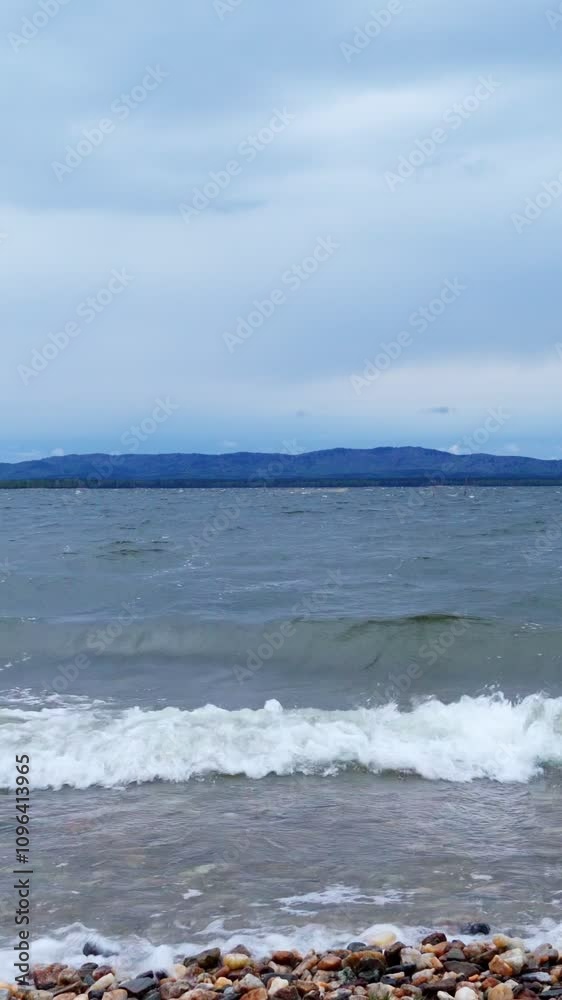 Coastal waves on the shore of the lake. The pebbly shore is washed by waves. Beauty is in nature. Vertical video, shorts. Coastal waves on the shore of the lake. The pebbly shore is washed by waves. Beauty is in nature. Vertical video, shorts.