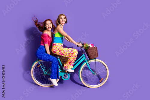 Full body portrait of two pretty lovely young girls ride bike empty space isolated on purple color background photo