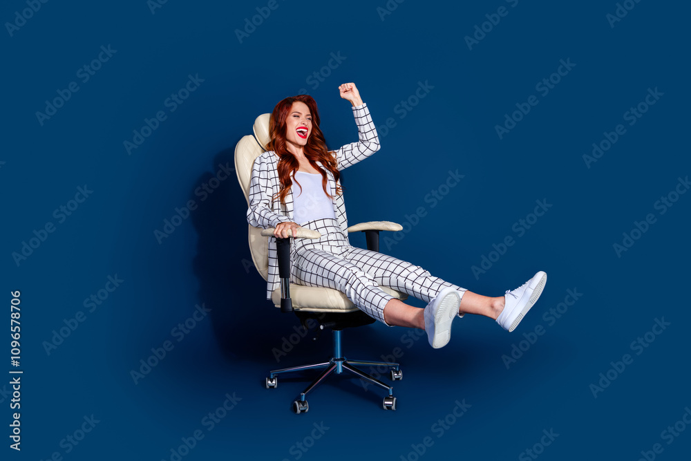 Young woman happily reclining in an office chair against a dark blue background, wearing a trendy checkered suit and white sneakers Young woman happily reclining in an office chair against a dark blue background, wearing a trendy checkered suit and white sneakers