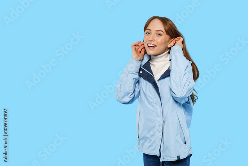 Young woman smiling in a light blue jacket against a solid blue background while playing with her hair