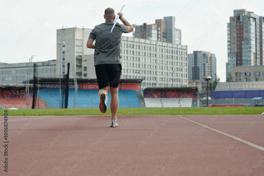 Obraz premium Athlete running at track while practicing javelin throw technique in city stadium with background of urban buildings during daytime offering dynamic sports action