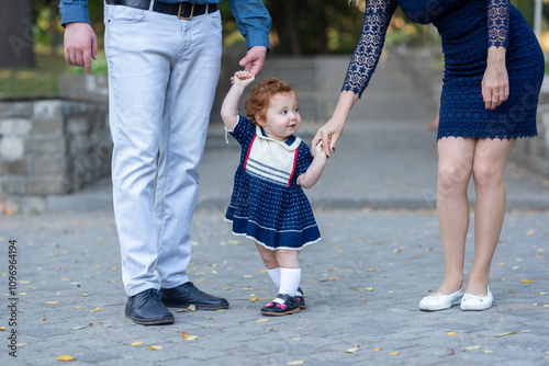 A cute curly-haired red-haired girl holds hands with her mom and dad and takes the first uncertain steps. Parenthood. Happiness  photo