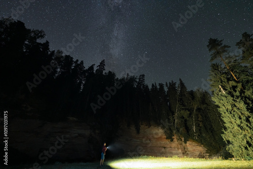 Night landscape astrophotography, man with flashlight hiking on the river bank near Suur Taevaskoja sandstone in autumn. Starry sky and milky way. Estonian nature.