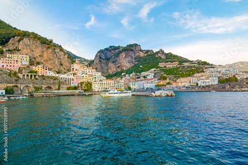 The photo captures a sunny view of Amalfi, showcasing the town's colorful buildings, steep cliffs, and vibrant coastline under a clear blue sky on a bright day.