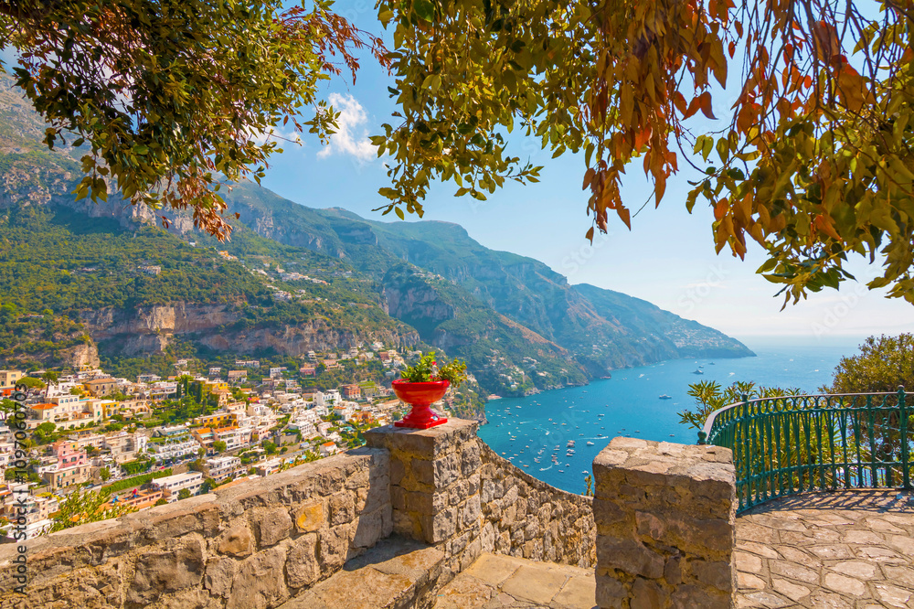 The photo captures a view of Positano, with its colorful, stacked houses cascading down the cliffs, narrow winding streets, and vibrant coastline set against the clear blue sea and sky