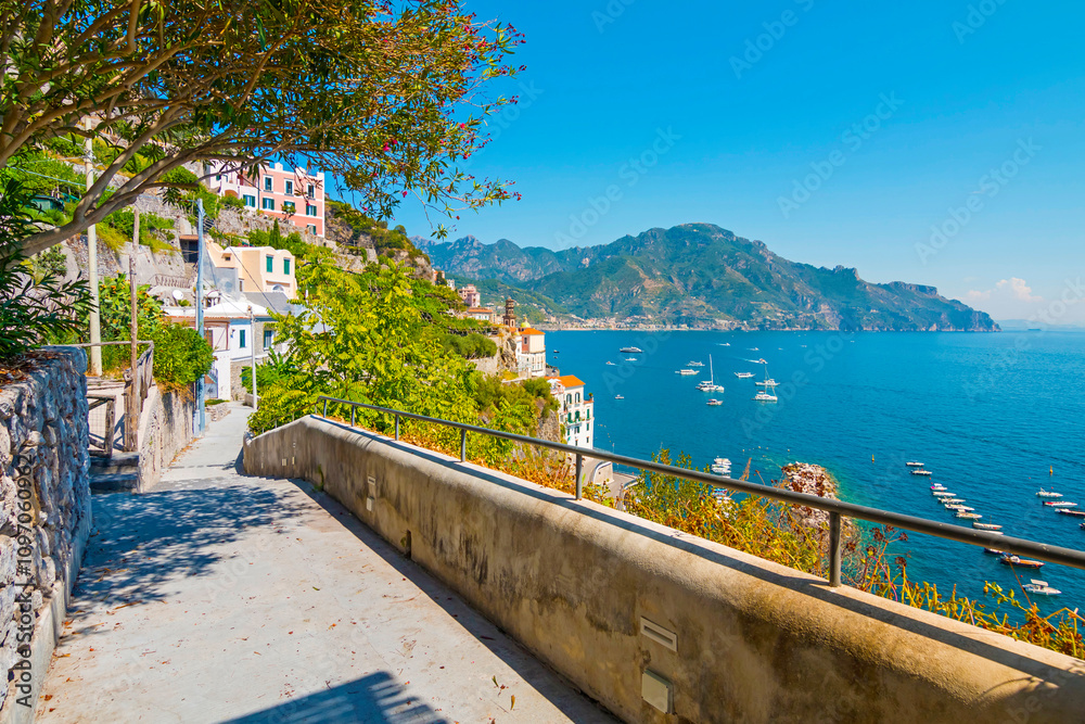 The photo captures a sunny view of Amalfi, showcasing the town's colorful buildings, steep cliffs, and vibrant coastline under a clear blue sky on a bright day.