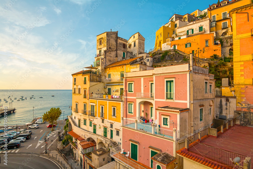 The photo captures a sunny view of Minori on the Amalfi Coast, showcasing its charming coastal buildings, sandy beach, and surrounding green hills under a clear blue sky.