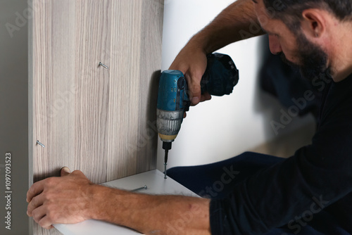 A man is assembling furniture using a cordless electric screwdriver. He is attaching a white shelf to a wooden panel, securing it with screws for stability. Image captures a precise moment of DIY work