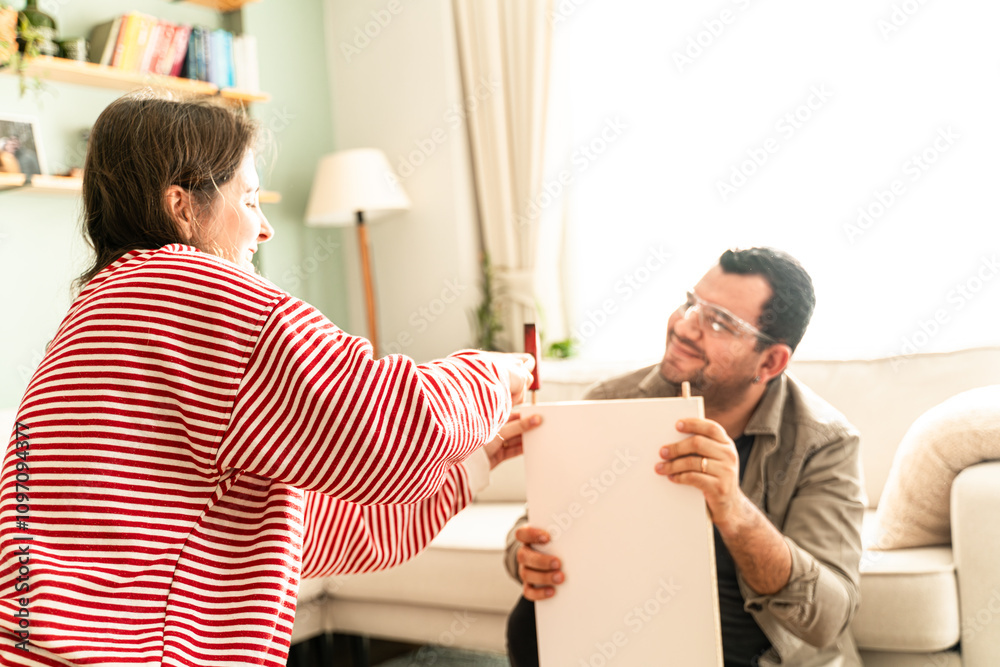 Fototapeta premium Happy Couple Assembles Furniture as a Team, Girl Reads Instructions and Boy Tightens Screw with a Drill. Moving into New Apartment, Couple Assemble Shelf.