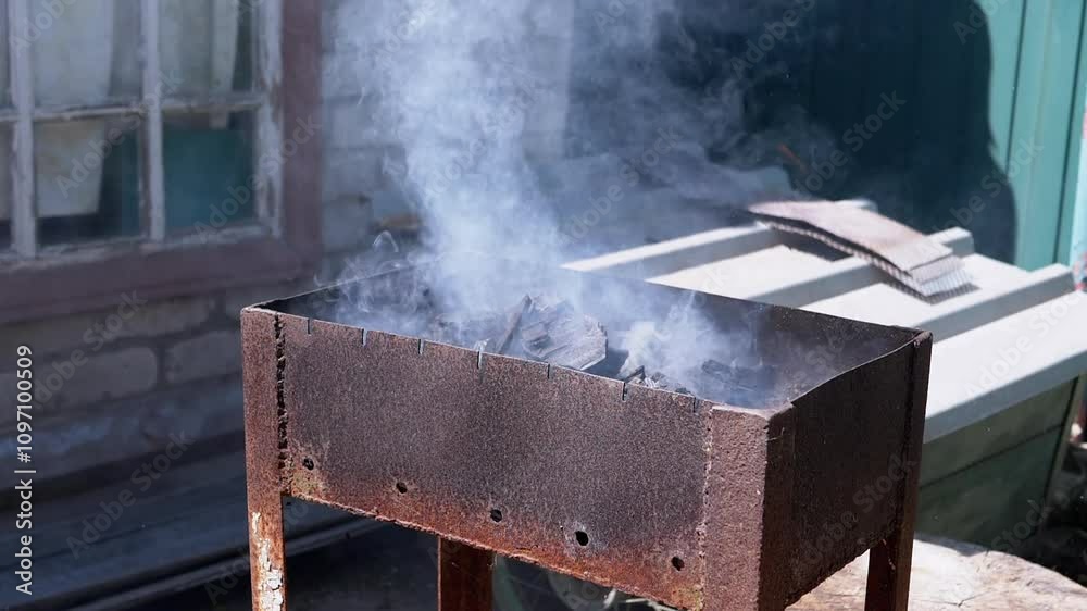 Close-up of a rusty brazier with smoldering coals, emitting white smoke, preparation for cooking meat outdoors. Perfect for projects related to BBQ, cooking, and summer holidays. Close-up of a rusty brazier with smoldering coals, emitting white smoke, preparation for cooking meat outdoors. Perfect for projects related to BBQ, cooking, and summer holidays.
