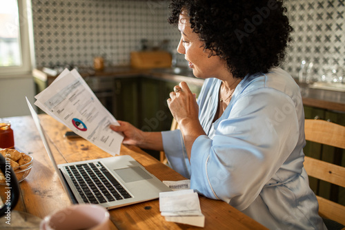 Focused woman reviewing bills and using laptop at home kitchen table photo