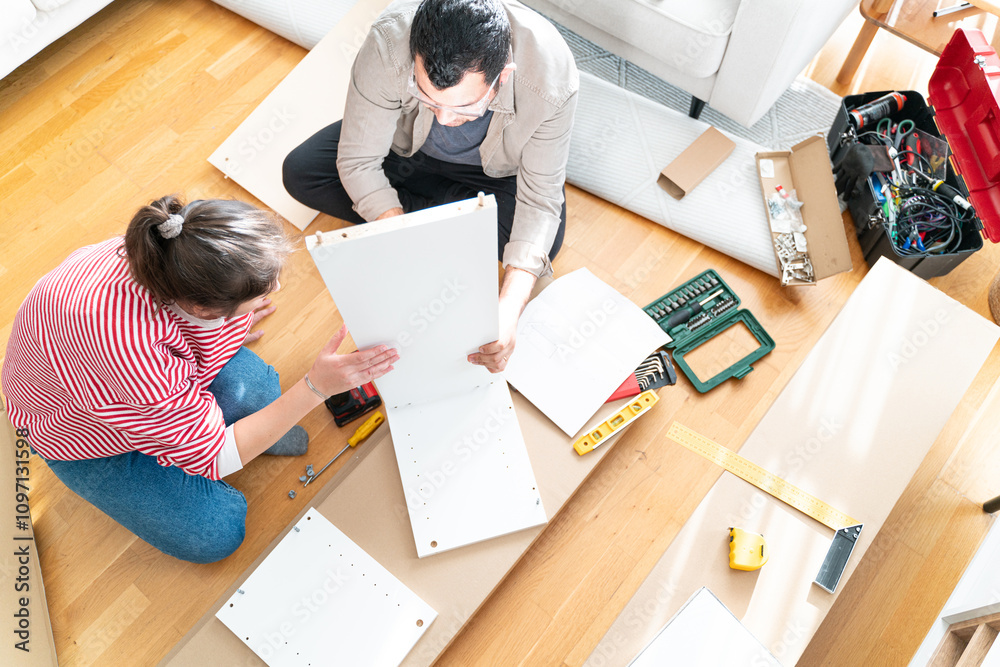 Happy Couple Assembles Furniture as a Team, Girl Reads Instructions and Boy Tightens Screw with a Drill. Moving into New Apartment, Couple Assemble Shelf. Happy Couple Assembles Furniture as a Team, Girl Reads Instructions and Boy Tightens Screw with a Drill. Moving into New Apartment, Couple Assemble Shelf.