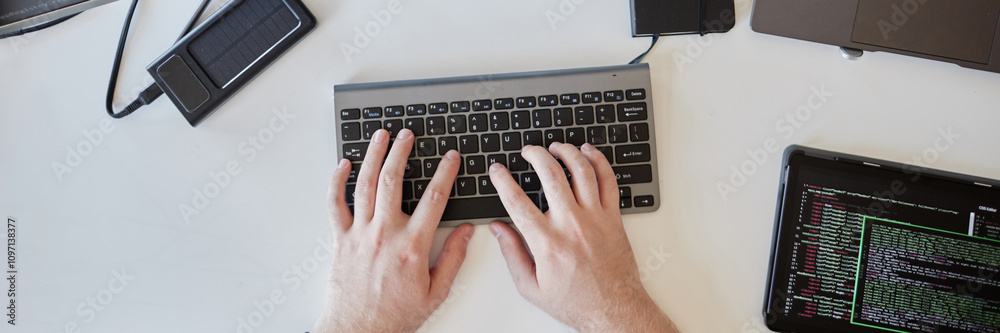 Hands typing on a wireless keyboard surrounded by various gadgets including a smartphone, power bank, and laptop suggesting tech-focused workspace Hands typing on a wireless keyboard surrounded by various gadgets including a smartphone, power bank, and laptop suggesting tech-focused workspace