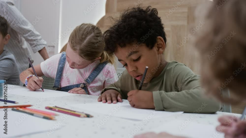 Handheld shot of group of diverse intercultural gen Alpha kids drawing on paper with pencils while sitting at table indoors Handheld shot of group of diverse intercultural gen Alpha kids drawing on paper with pencils while sitting at table indoors