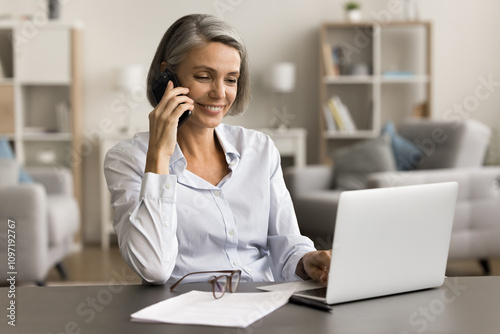 Happy positive middle aged business woman talking on cell at workplace table with laptop, speaking on mobile phone, using wireless technology for working from home, smiling, enjoying conversation