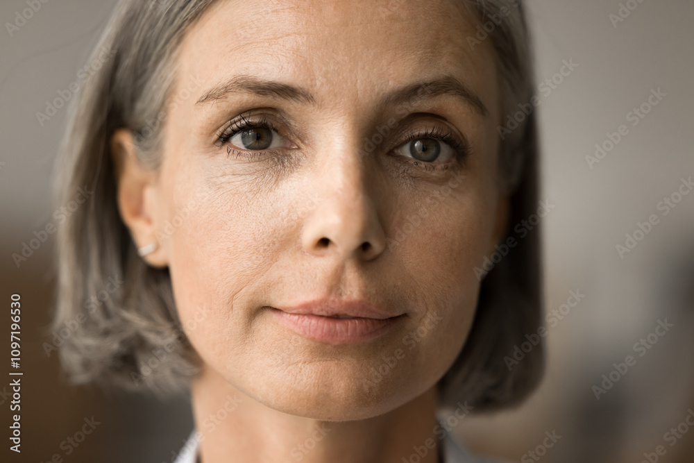 Fototapeta premium Pretty face of mature 50s grey haired woman looking at camera with positive serious calm expression. Senior lady with natural makeup promoting elderly age beauty care