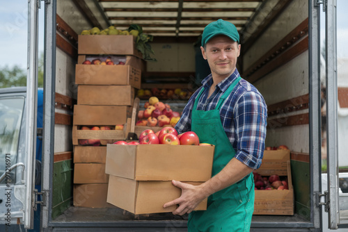 Confident delivery worker carrying fresh apples in a cardboard box from a truck for organic farming and healthy food lifestyle promotion