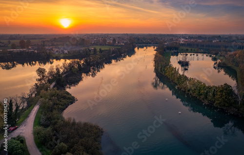 A vibrant sunset casts golden hues over the Sile river, reflecting on its calm waters. Lush greenery and winding paths line the riverbanks, creating a serene landscape in Casier, Veneto.