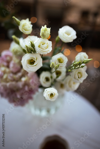 Bouquet of ranunculus blooms in a vase standing on a white table with cup of coffee on a french balcony in the evening with Christmas lights on its background