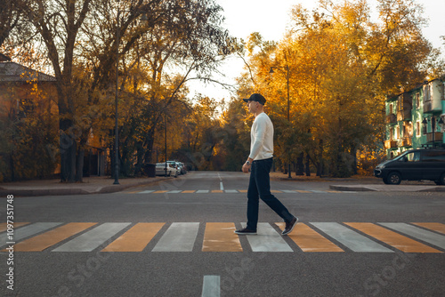 Young Man Walking Across Urban Crosswalk During Golden Autumn Sunset, Peaceful City Street Scene with Fall Foliage and Residential Neighborhood Setting