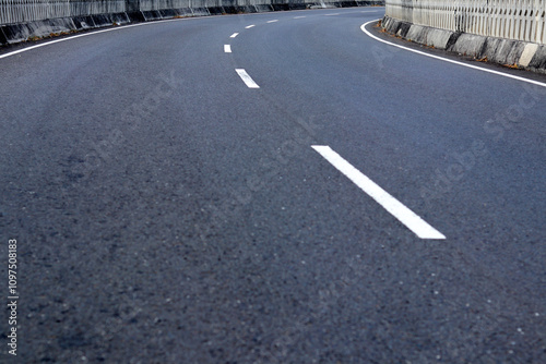 A smooth, winding asphalt road curves gently, marked by crisp white lines, inviting drivers to explore the journey ahead amidst a serene urban landscape. photo