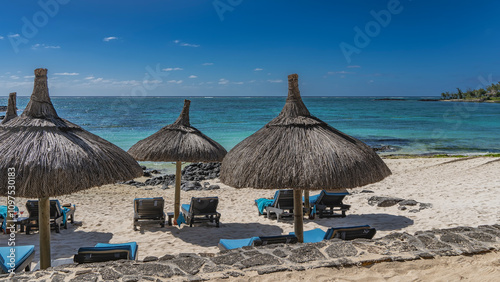 Sun loungers with soft mattresses on the sandy beach in the shade of straw umbrellas. The turquoise ocean is calm. Black volcanic boulders by the water. Blue sky, clouds. Mauritius Belle Mare