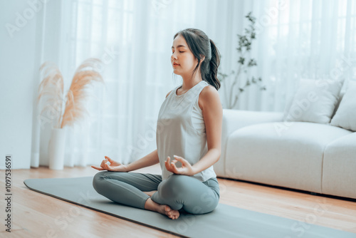 Young Asian woman meditating in lotus position at home, sitting on the floor in fitness clothing. Breathing workout to achieve relaxation, peace and mindfulness photo