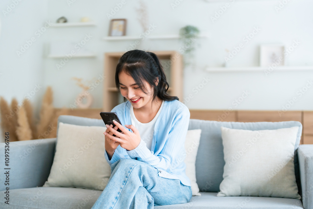 Smiling pretty young Asian woman sitting on cozy couch, using funny mobile apps in living room. Woman at Home, Doing Online Shopping, Messaging Friends, Posting on Social Media Smiling pretty young Asian woman sitting on cozy couch, using funny mobile apps in living room. Woman at Home, Doing Online Shopping, Messaging Friends, Posting on Social Media