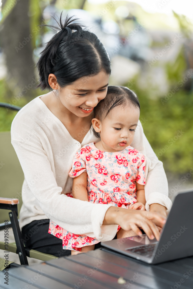 A young mother sits at a table with her smiling daughter on her lap. She types on a laptop while the cheerful little girl looks at the camera, radiating happiness. A young mother sits at a table with her smiling daughter on her lap. She types on a laptop while the cheerful little girl looks at the camera, radiating happiness.
