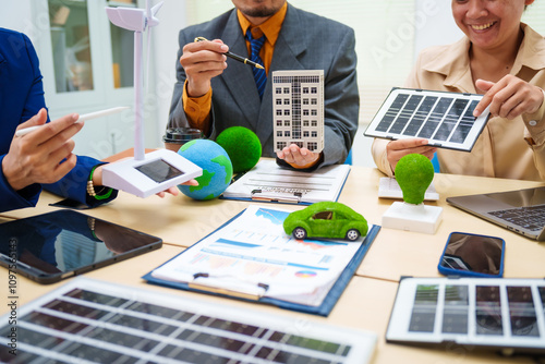 Asian business professionals in collaborate at desk with building models, planning sustainable projects solar panels, electric cars, wind turbines, eco-friendly practices and clean energy solutions. photo