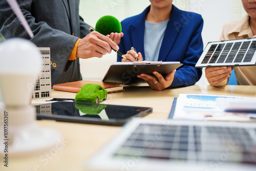 Asian business professionals in collaborate at desk with building models, planning sustainable projects solar panels, electric cars, wind turbines, eco-friendly practices and clean energy solutions. photo