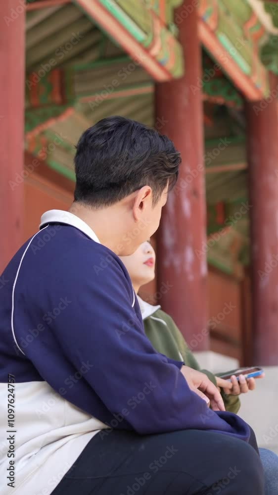 A Korean couple, a man in his 30s and a woman in her 20s, wearing tracksuits, sit together and talk in a historic park in Seoul, South Korea. Vertical video. A Korean couple, a man in his 30s and a woman in her 20s, wearing tracksuits, sit together and talk in a historic park in Seoul, South Korea. Vertical video.