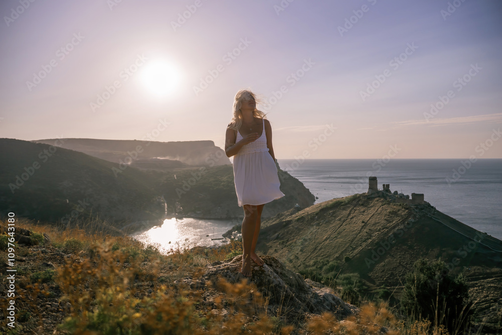 A blonde woman stands on a hill overlooking the ocean. She is wearing a white dress and she is enjoying the view. A blonde woman stands on a hill overlooking the ocean. She is wearing a white dress and she is enjoying the view.