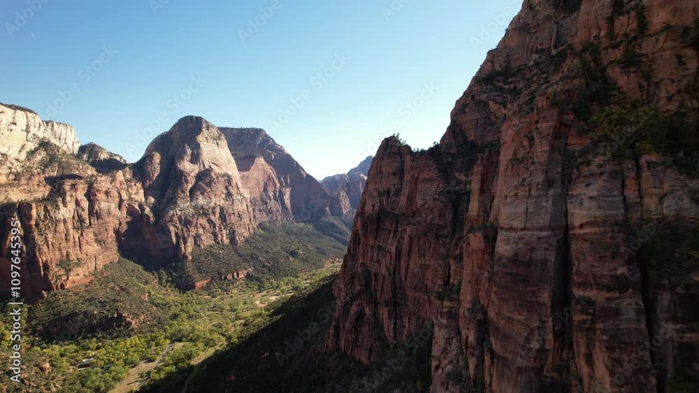 Aerial shot showcasing the dramatic red cliffs of Zion Canyon with lush greenery below and distant peaks under a clear sky. Aerial shot showcasing the dramatic red cliffs of Zion Canyon with lush greenery below and distant peaks under a clear sky.