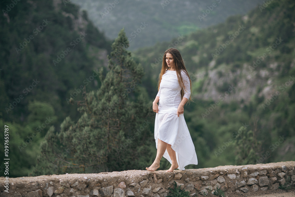 Naklejka premium A woman in a white dress is walking on a stone wall. The image has a serene and peaceful mood, as the woman is alone and surrounded by nature.
