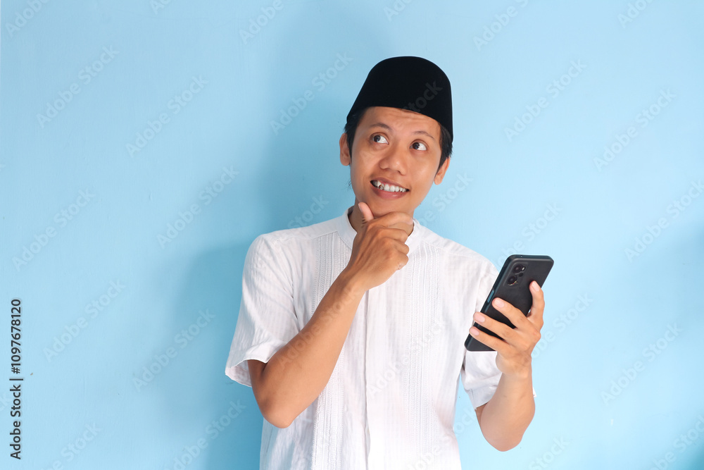 Young asian muslim man smiling holding his mobile phone while looking side with touching his chin Young asian muslim man smiling holding his mobile phone while looking side with touching his chin