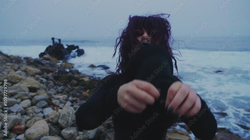 A masked figure in black stands at the seashore and pulls off a torn black mask, revealing a young Goth girl. Mysterious masked woman at the rocky seacoast.