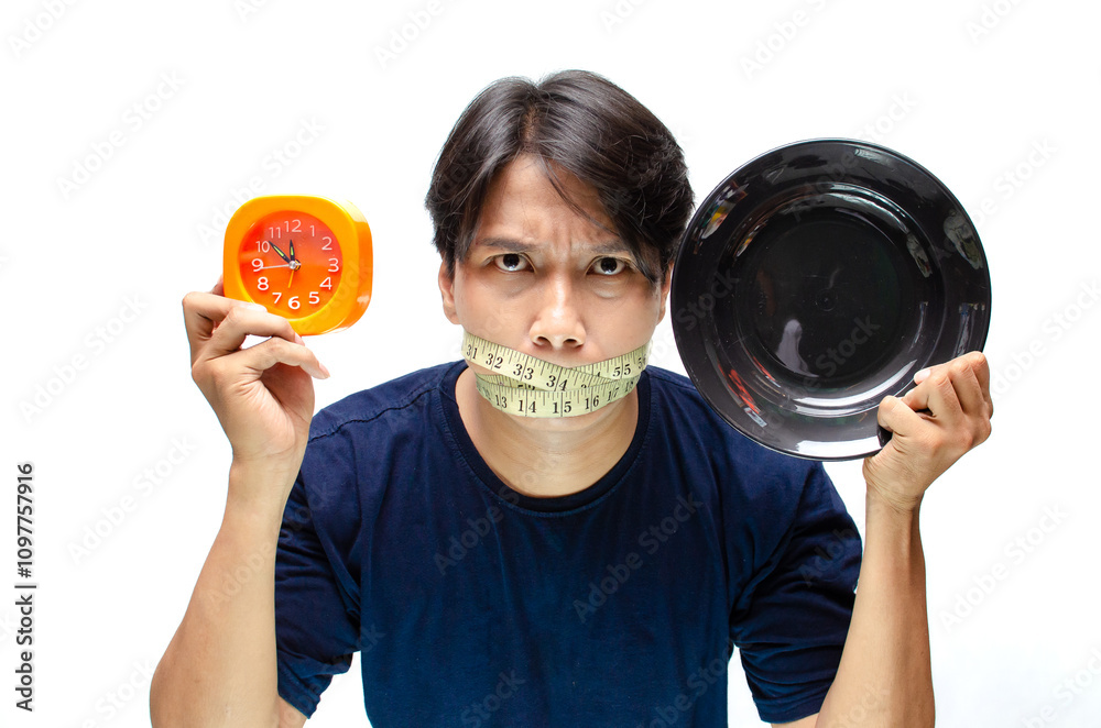 A frustated young Asian man on diet covered his mouth using measurement tape while holding an empty plate and clock as a concept of waiting time to break fasting. A frustated young Asian man on diet covered his mouth using measurement tape while holding an empty plate and clock as a concept of waiting time to break fasting.