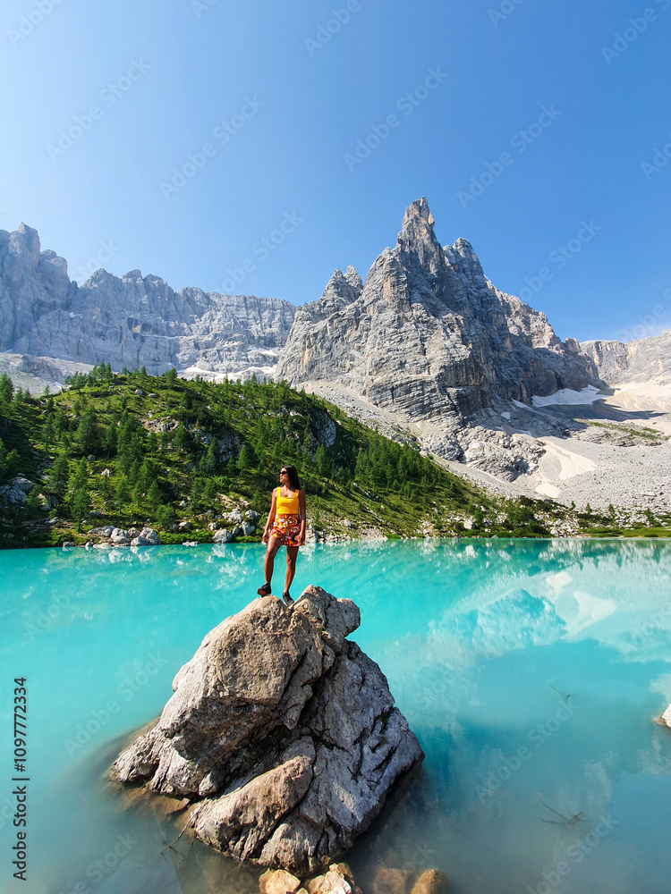A person stands triumphantly on a rock by a stunning turquoise lake Lago Di Sorapis Dolomites Italy A person stands triumphantly on a rock by a stunning turquoise lake Lago Di Sorapis Dolomites Italy