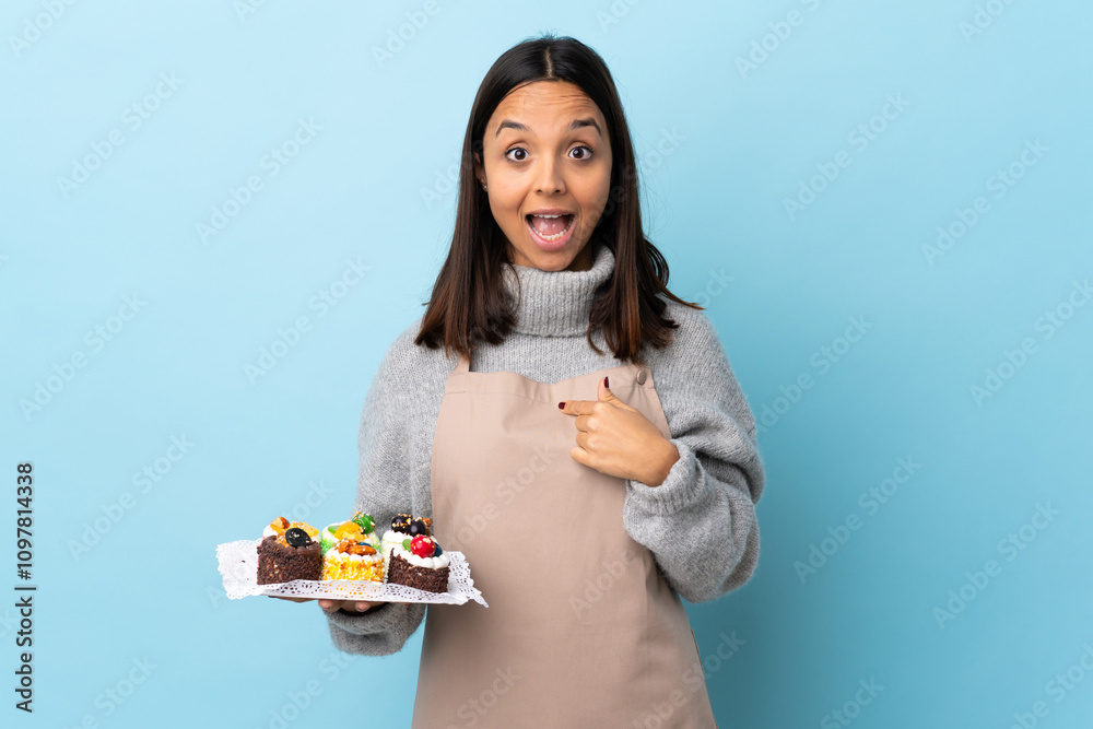 Pastry chef holding a big cake over isolated blue background with surprise facial expression. Pastry chef holding a big cake over isolated blue background with surprise facial expression.
