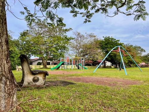 A vibrant playground with swings, slides, and seesaws in a lush residential park, surrounded by shady trees and clean walking paths.