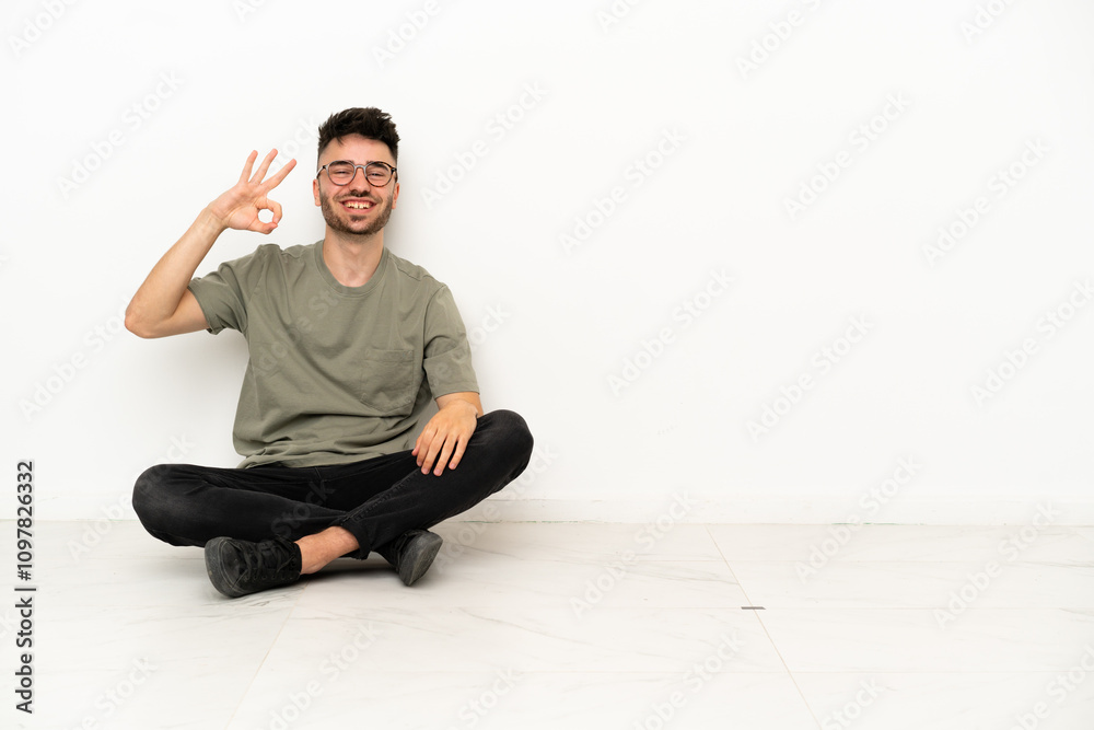 Young caucasian man sitting on the floor isolated on white background showing ok sign with fingers Young caucasian man sitting on the floor isolated on white background showing ok sign with fingers