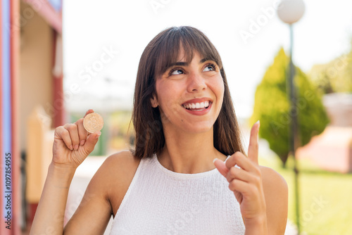 Young woman holding a Bitcoin at outdoors intending to realizes the solution while lifting a finger up