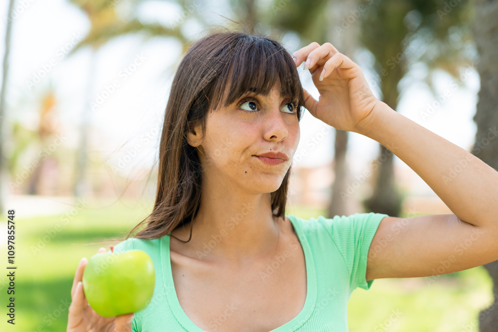 Young woman with an apple at outdoors having doubts and with confuse face expression Young woman with an apple at outdoors having doubts and with confuse face expression