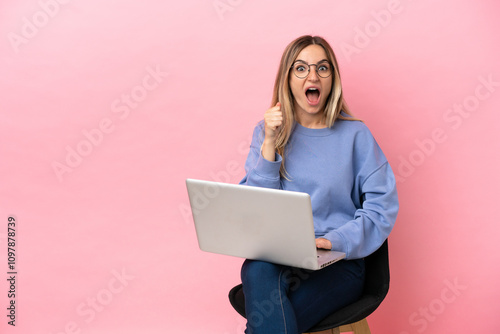 Young woman sitting on a chair with laptop over isolated pink background celebrating a victory in winner position