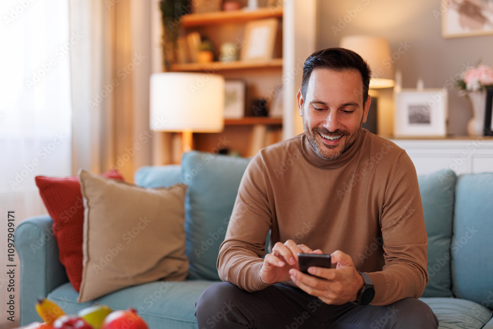 Obraz premium Portrait of handsome man smiling and scrolling apps over smart phone while sitting on sofa at home
