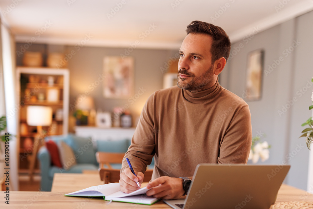 Serious businessman looking away thoughtfully while writing notes and working over laptop at desk