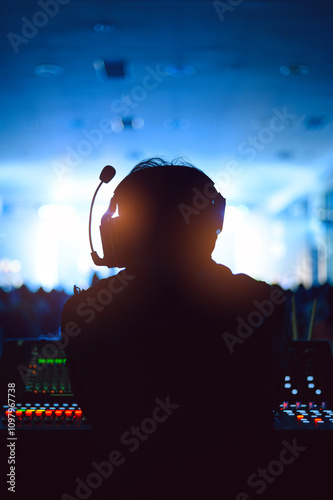 Silhouette of a sound technician wearing a headset, working at a control panel during a live event. The background features blue stage lighting and a blurred audience, creating a vibrant atmosphere.