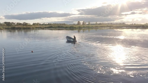 A person rows a small boat across a wide lake. Sunlight reflects on the water, with clouds in the sky. Trees line the horizon, and a buoy floats nearby. photo